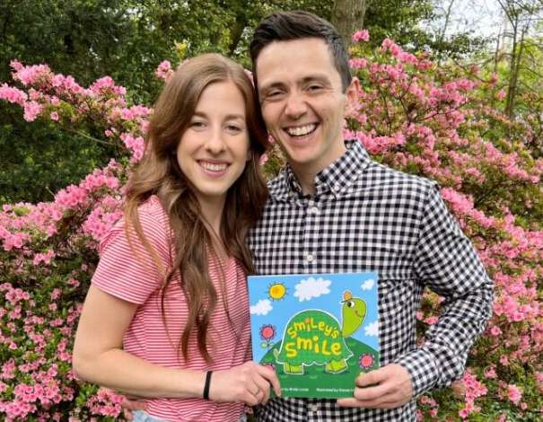 Brady holding his book “Smiley’s Smile” along with his wife Julia as they stand in front of a blooming pink floral tree.
