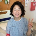 Caryn smiling and giving a peace sign while wearing a hospital gown sitting on her hospital bed.